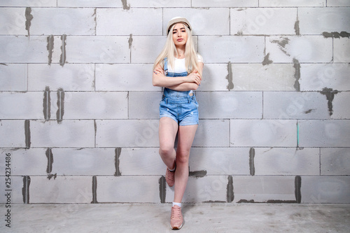 Cute girl designer foreman blonde in construction helmet in denim overalls standing near gray wall of Cibit in house under construction. Сoncept repair, female labor, photo session repair