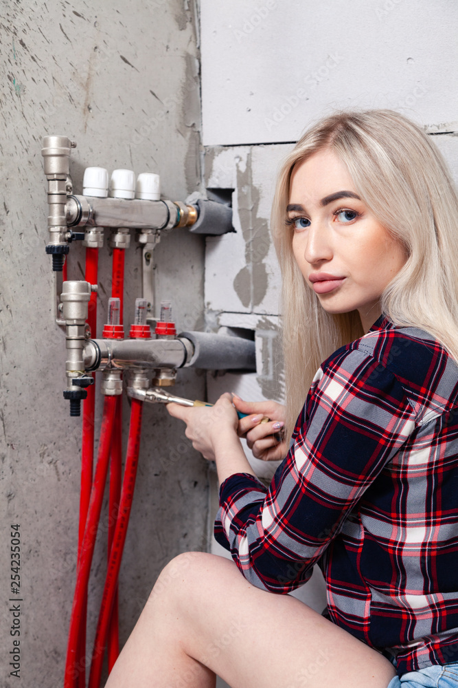 Blonde girl foreman tightens the screw nuts with wrench on control system of warm water floor in house under construction. Concept professional worker, female labor, photo model, shoot, repair