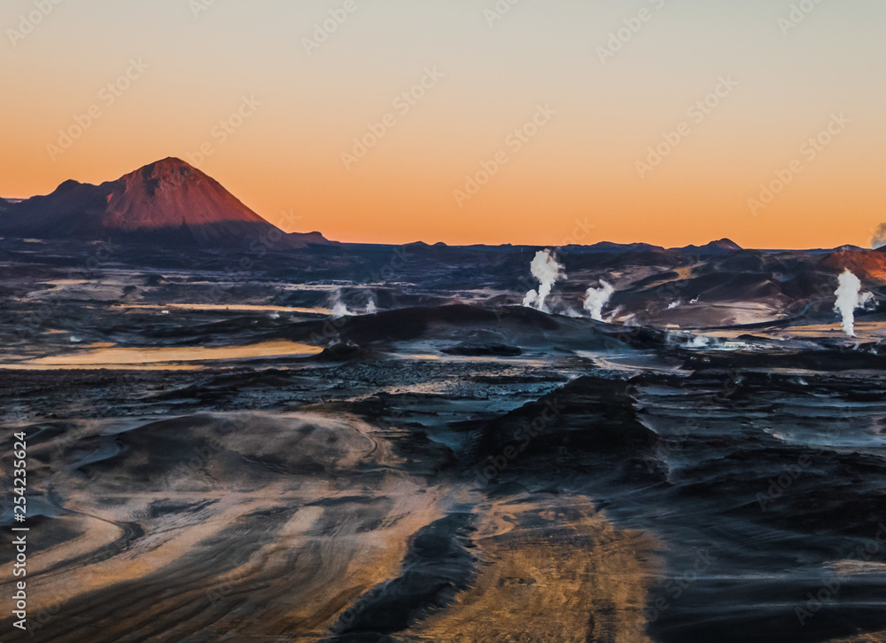 A volcanic, geothermal landscape captured during the sunrise. First beams of the sun reach the top parts of volcano. Smoke coming out of hot springs. Volcanic lava fields all around. Cosmic and alien
