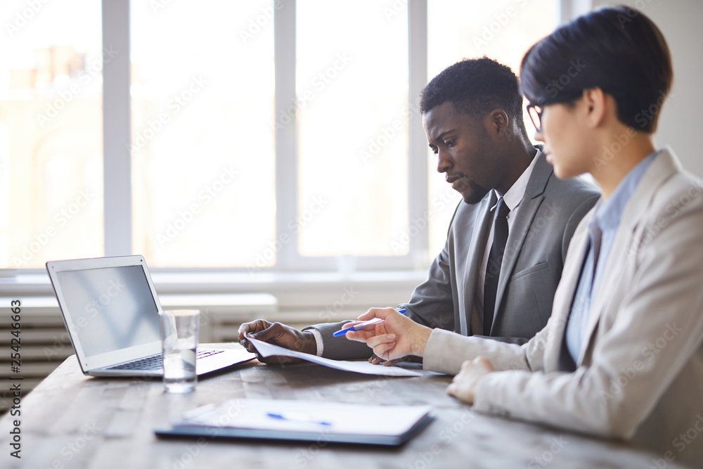 Two young intercultural business employees in formalwear sitting by desk in office and discussing papers Two young intercultural business employees in formalwear sitting by desk in office and discussing papers