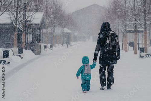 Back view of affectionate father and little male child hold hands, walk along snowy street during heavy snowfall in winter, enjoy recreation time, go home, dressed in warm clothes, carry rucksack