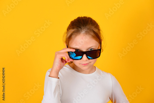 A girl in sunglasses holds them and looks into the camera, on a yellow background in the studio.