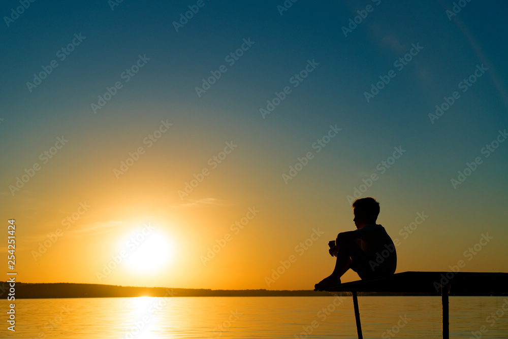 Small boy sitting alone on the footbridge holding his legs and dreaming at sunset. Little kid watches the sun setting over the river.