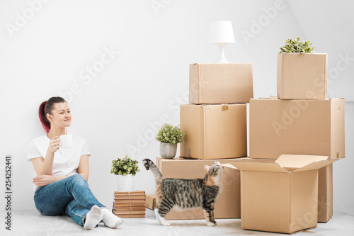 A young beautiful brunette girl in a white T-shirt is sitting on the floor of a bright room and making calls on her smartphone. Around cardboard boxes and a cat. © spaskov