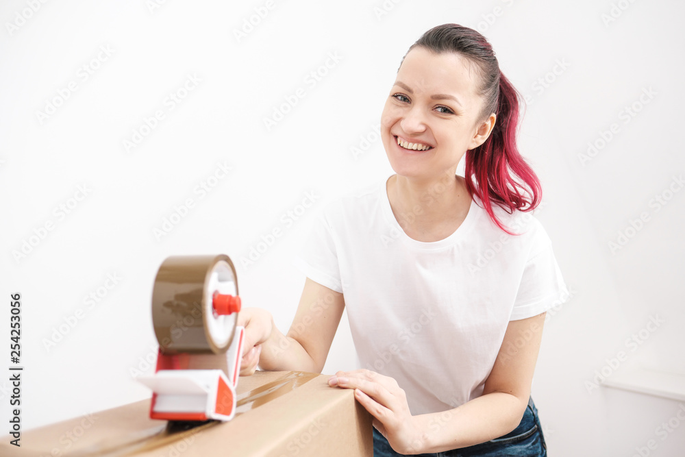 Young beautiful brunette girl in a white t-shirt packs cardboard boxes with a dispenser and adhesive scotch adhesive tape. The concept of moving to a new home. Young beautiful brunette girl in a white t-shirt packs cardboard boxes with a dispenser and adhesive scotch adhesive tape. The concept of moving to a new home.