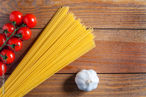 Spaghetti, cherry tomatoes and bulb of garlic on a brown wooden table. Tomatoes to the left of spaghetti, garlic to the right. Top view.