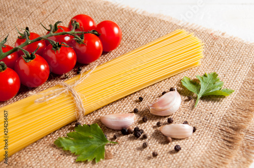 Bunch of raw spaghetti, cherry tomatoes, peppers, garlic cloves and two parsley leaves on burlap on a white wooden background. Close-up.