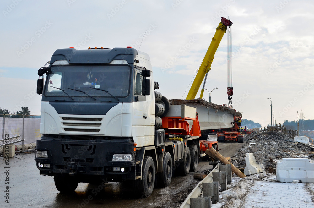 A crane unloads a long concrete slab from a truck at a construction site. Workers unload reinforced concrete panels