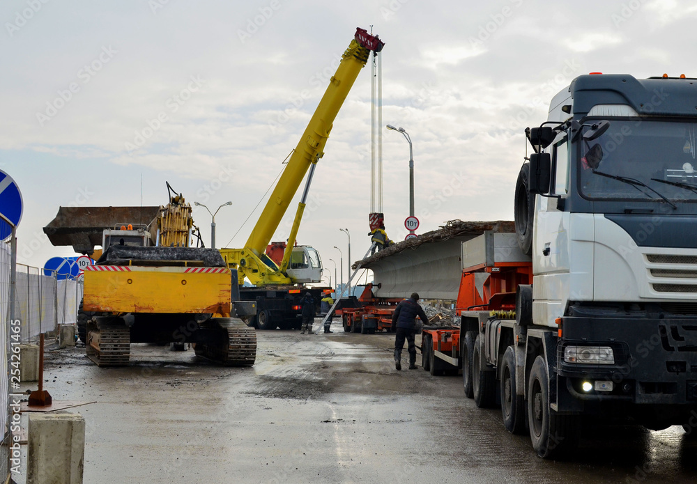 A crane unloads a long concrete slab from a truck at a construction site. Workers unload reinforced concrete panels