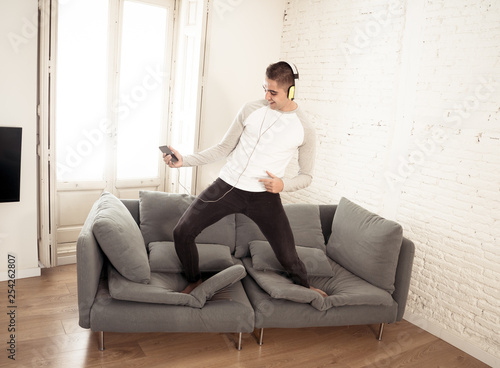 Young man in his twenties with headphones listening to music and dancing on sofa at home
