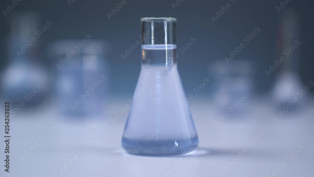 On the table is a medical flask with liquid. The camera smoothly leaves the flask. Gray wall on background. Flasks in defocus on the gray background. On the table is a medical flask with liquid. The camera smoothly leaves the flask. Gray wall on background. Flasks in defocus on the gray background.
