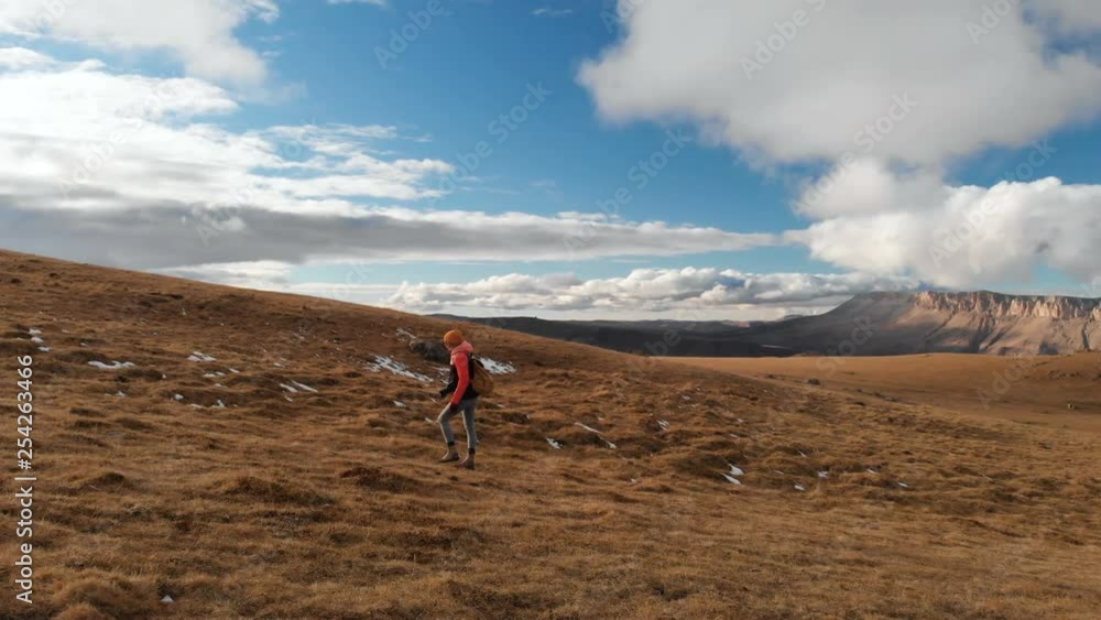 Air view. Girl traveler with a camera on her neck walking along the slope with yellow grass against the backdrop of mountains and a plateau with clouds at sunset Air view. Girl traveler with a camera on her neck walking along the slope with yellow grass against the backdrop of mountains and a plateau with clouds at sunset