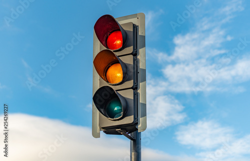 An English traffic light sits isolated against a bright blue sky with the red, orange and green lights