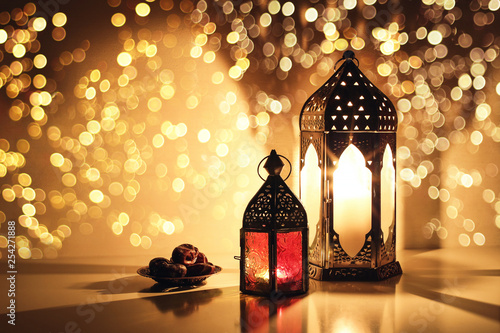 Ornamental Arabic lanterns with burning candles. Glittering golden bokeh lights. Plate with date fruit on the table. Greeting card for Muslim holiday Ramadan Kareem. Iftar dinner background.