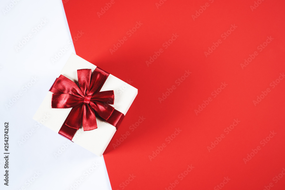 Beautiful present box tied up with shiny satin ribbon on contrast red and white background with copy space for text. Preparation for holidays and celebration concept. Beautiful present box tied up with shiny satin ribbon on contrast red and white background with copy space for text. Preparation for holidays and celebration concept.