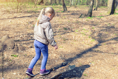 Cute little caucasian blond girl walking on wooden log in forest. Adorable child having fun during walk in park in bright sunny day