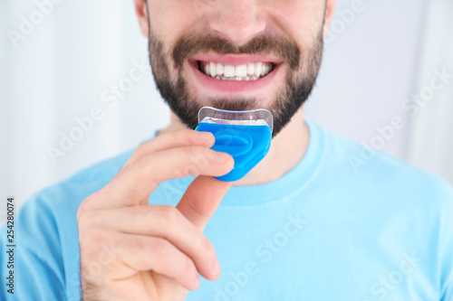 Young man using teeth whitening device on light background, closeup