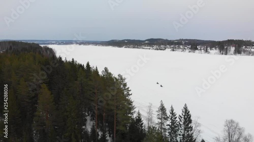 Aerial video flying over a frozen lake and the woods in northern Sweden with two snowmobiles driving by photo