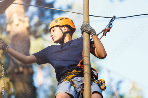 Fototapeta Sporty, young, cute boy in white t shirt spends his time in adventure rope park in helmet and safe equipment in the park in the summer