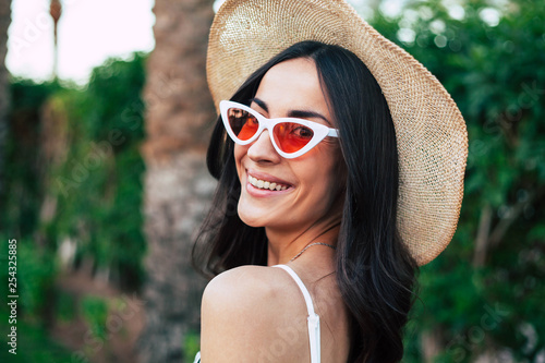 Travel bug. Gorgeous girl with long dark hair, brilliant smile and hazel eyes wearing camel hat and cat shaped sunglasses in front of deep green bushes and a palm tree.