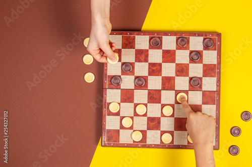Child hands playing checkers on checker board game over yellow and blue background, top view