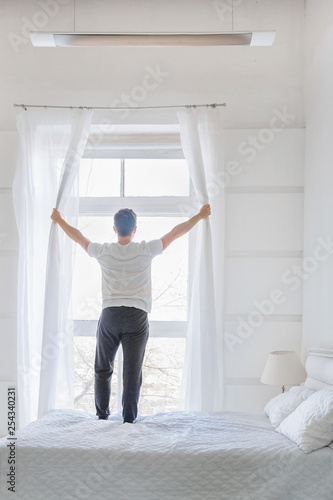 Young man stand near window in modern white bedroom. Happy man open curtains up in the morning. Rear view