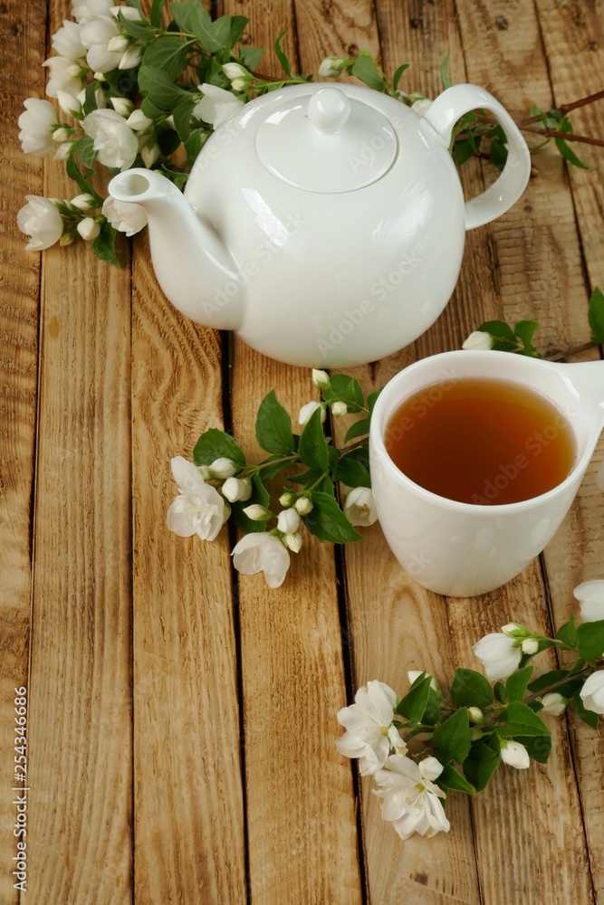 Jasmine tea. tea with jasmine extract, white teapot and white mug with tea, blooming sprigs of jasmine on a wooden brown background.Herbal organic wholesome tea Jasmine tea. tea with jasmine extract, white teapot and white mug with tea, blooming sprigs of jasmine on a wooden brown background.Herbal organic wholesome tea
