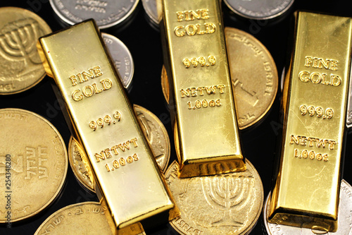 A macro image of miscellaneous coins from Israel with three small gold ingots on a reflective black background
