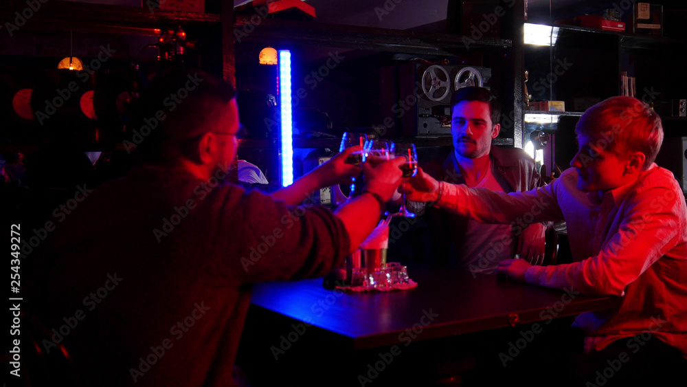 Bar with neon lighting. Group of friends sitting by the table and drinking alcohol. Cheers