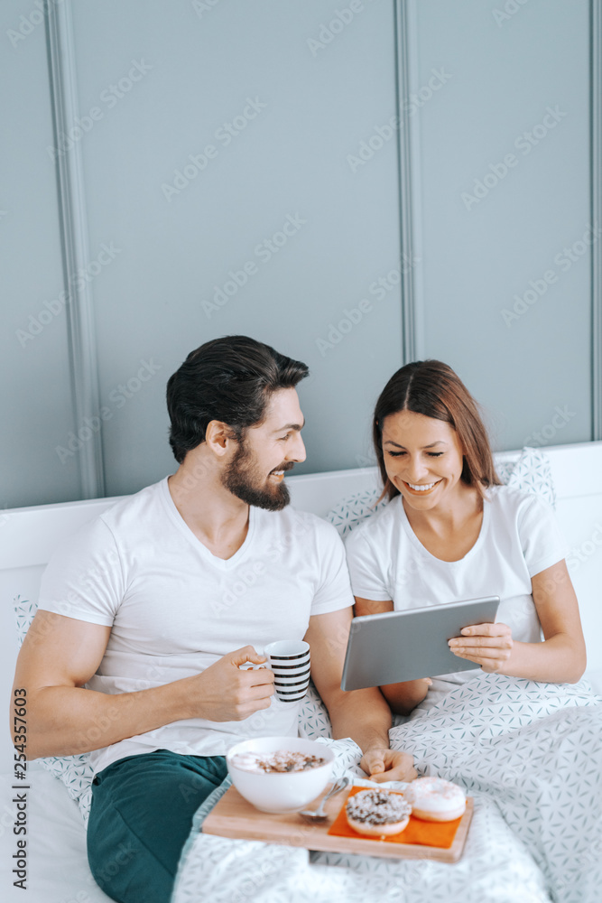 Young happy Caucasian couple in love having breakfast in bed and watching videos on tablet.