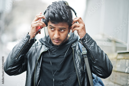 Stylish and casual asian man in black leather jacket, headphones with red mobile phone at hands posed on street and listening music.