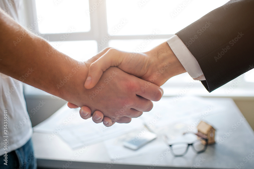 Close up cut view shaking hands of two men. Table with apartment plan and window on back. Bright background.