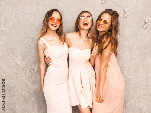 Three young beautiful smiling hipster girls in trendy summer light pink dresses. Sexy carefree women posing near gray wall. Positive models in round sunglasses having fun