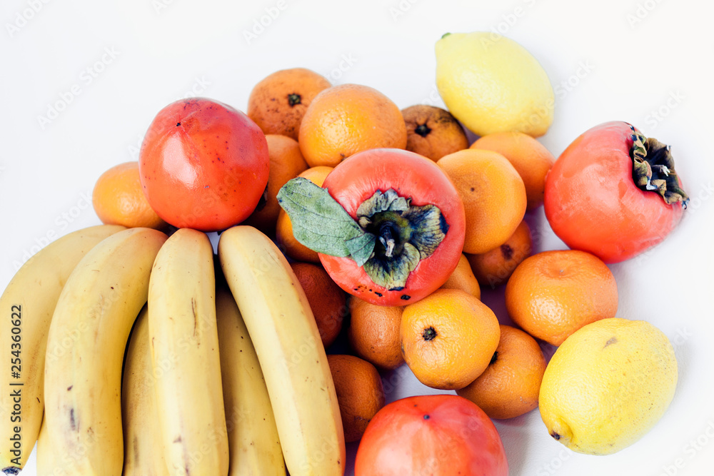 A selection of arranged different fresh fruits of bananas, mandarins, persimmons and lemons on white background close up. A selection of arranged different fresh fruits of bananas, mandarins, persimmons and lemons on white background close up.