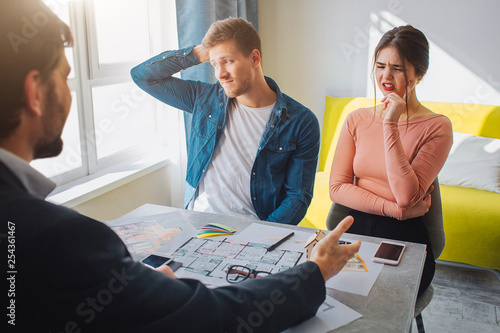 Couple buy or rent apartment together. Confused suspicious young man and woman have doubts. They sit in front of realtor. He reach hand and point forward.