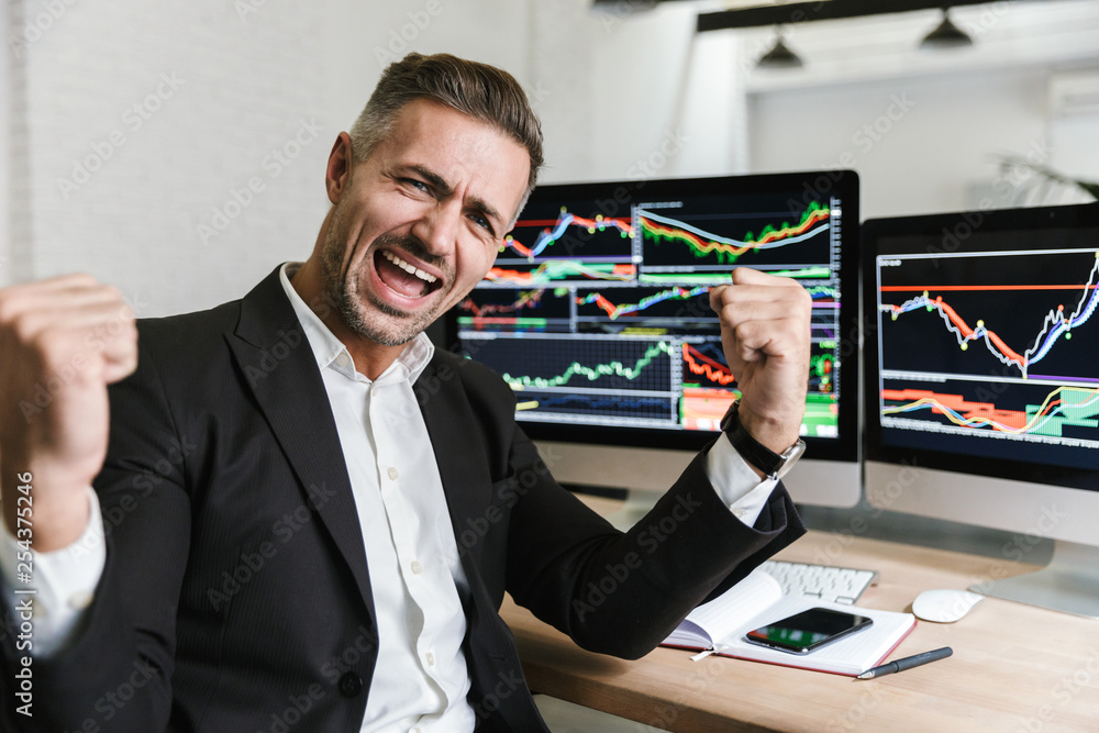 Photo of ecstatic businessman rejoicing while working in office with digital graphics and charts Photo of ecstatic businessman rejoicing while working in office with digital graphics and charts