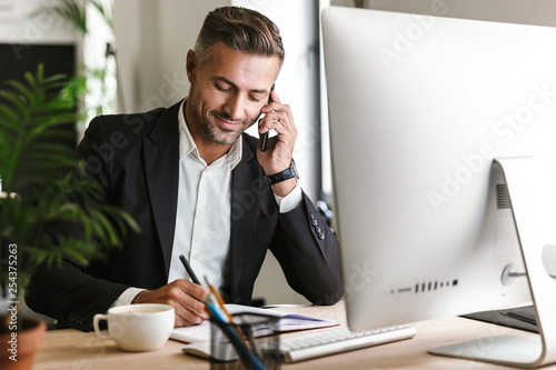 Image of handsome businessman talking on cell phone while working on computer in office