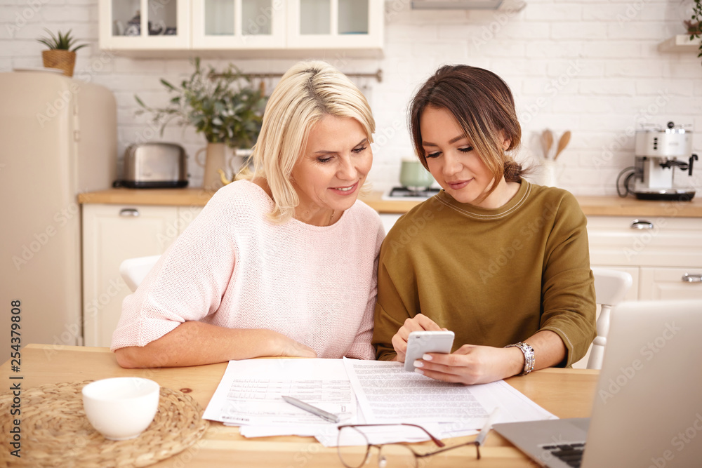 Indoor image of beautiful dark haired young woman holding mobile phone, showing her attractive middle aged mother how to pay mortgage or apartment rent online using application on electronic gadget Indoor image of beautiful dark haired young woman holding mobile phone, showing her attractive middle aged mother how to pay mortgage or apartment rent online using application on electronic gadget