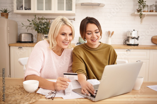 Happy mature blonde female holding credit card sitting at kitchen table with her cute young daughter, shopping online, buying new clothes, booking accomodation or airplane tickets, planning vacations