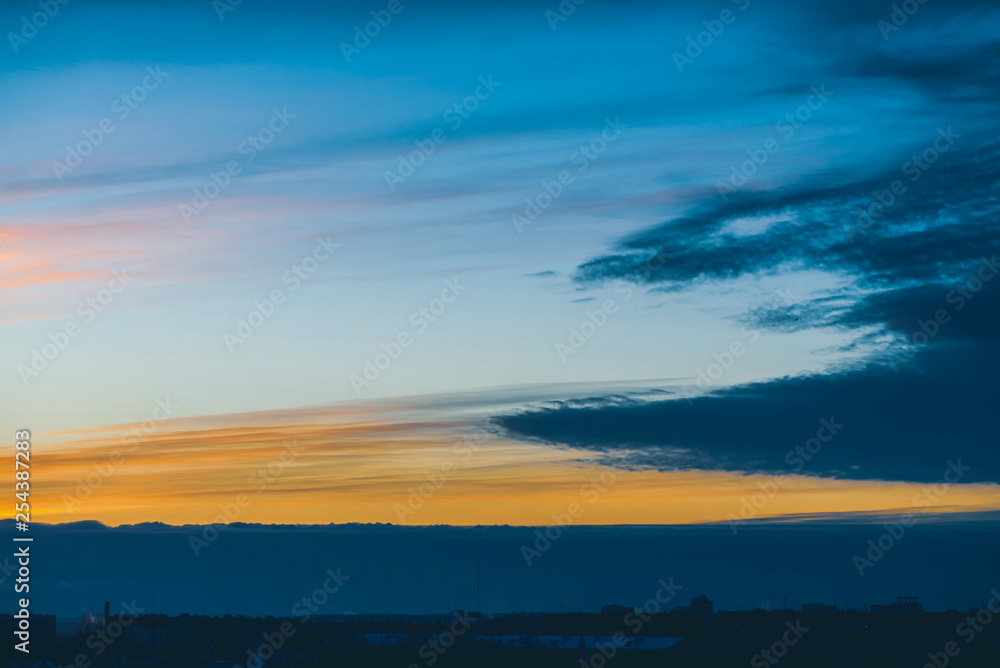 Cityscape with wonderful varicolored vivid dawn. Amazing dramatic blue cloud sky above dark silhouettes of city building roofs. Atmospheric background of orange sunrise in overcast weather. Copy space