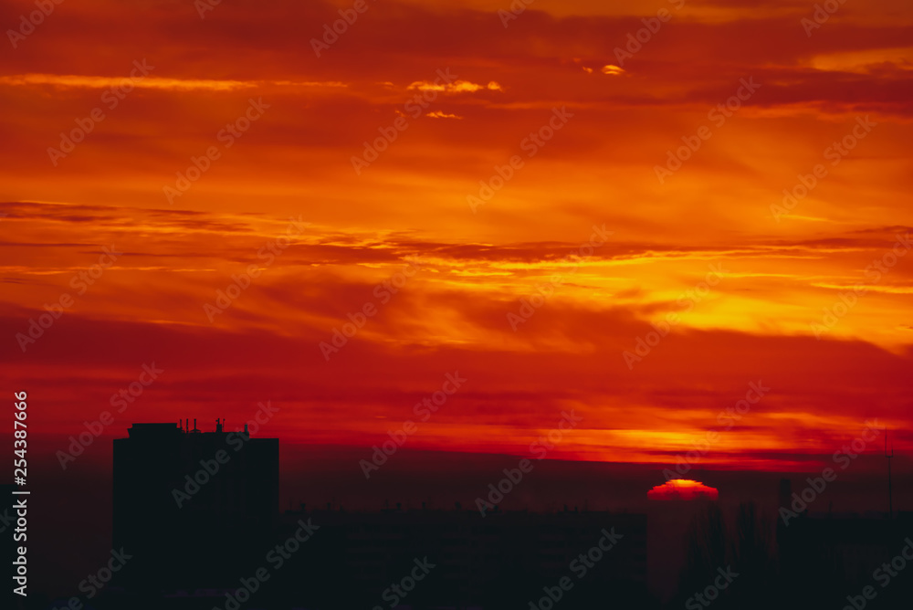 Sun rises over horizon. Fiery sunny circle. Cityscape with vivid red dawn. Amazing warm dramatic cloudy sky above dark silhouettes of city building roofs. Atmospheric background of sunrise. Copy space