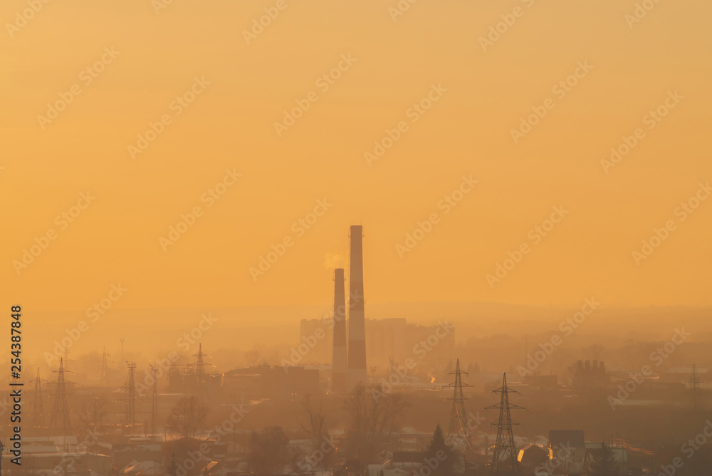 Smog among silhouettes of buildings on sunrise. Smokestack in dawn sky. Environmental pollution on sunset. Harmful fumes from stack above city. Mist urban background with warm orange yellow sky. Smog among silhouettes of buildings on sunrise. Smokestack in dawn sky. Environmental pollution on sunset. Harmful fumes from stack above city. Mist urban background with warm orange yellow sky.