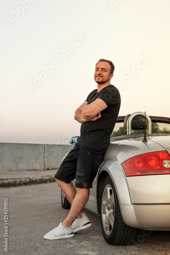 Gorgeous sexy man on a vacation car trip in summer day. Luxury grey sport car. Man in casual outfit. Sunset
