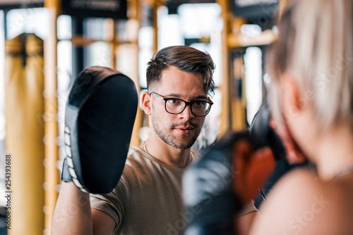 Male trainer exercising with young sportswoman in boxing gloves at gym.