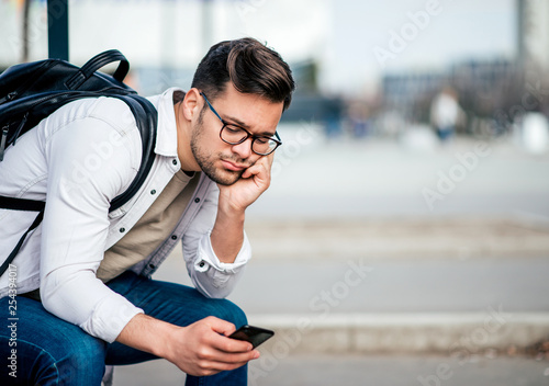 Casual bored young man with backcpack sitting on bench and waiting for bus, using smartphone. photo