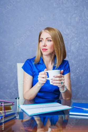 A woman in a blue dress sits at a table with notepads and a diary and holds a mug of tea. Office theme.