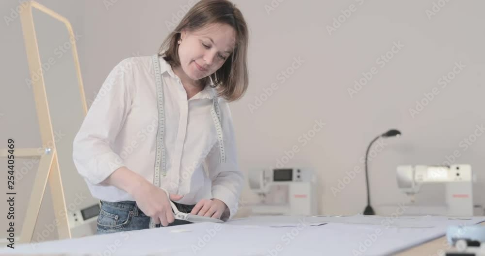 Slow motion video of the fashion designer which works at a table with curves and a pattern, she are cutting fabric, around lies scissors, centimeter, sew machine and dummy on the background Slow motion video of the fashion designer which works at a table with curves and a pattern, she are cutting fabric, around lies scissors, centimeter, sew machine and dummy on the background