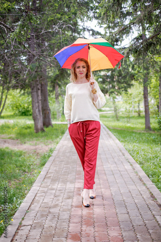 A woman in a light jumper and red trousers outside with a colorful rainbow umbrella