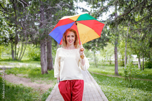 A woman in a light jumper and red trousers outside with a colorful rainbow umbrella