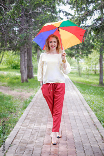 A woman in a light jumper and red trousers outside with a colorful rainbow umbrella